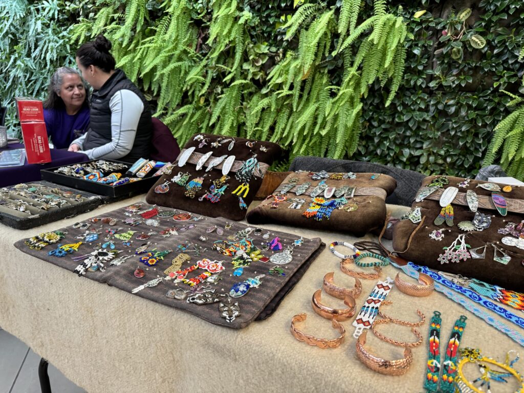 A table display featuring an array of handcrafted jewelry and accessories, including beaded bracelets, earrings, pendants, and copper bracelets. The items are arranged on fabric and wooden stands, showcasing vibrant colours and intricate designs. In the background, two women are seated and engaged in conversation. In the background is a lush green living plant wall.